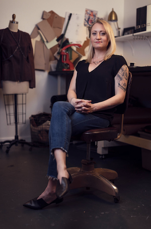 light skinned woman with blonde hair and tattoos sits in a chair in a fashion design studio
