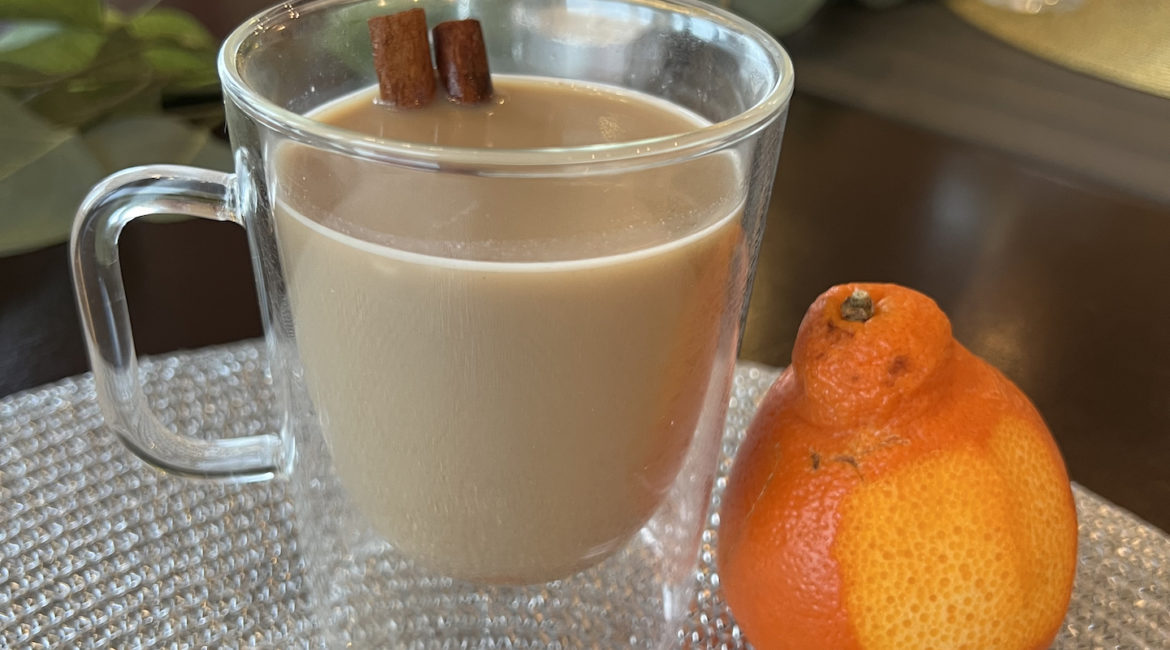 A clear glass mug with a handle sites on a silver placemat. The mug is filled with a chai tea mocktail and has a cinnamon stick poking out of the top of the beverage as a stir stick. An orange with a small amount of the peel removed is placed next to the mug.