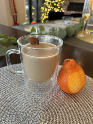 A clear glass mug with a handle sites on a silver placemat. The mug is filled with a chai tea mocktail and has a cinnamon stick poking out of the top of the beverage as a stir stick. An orange with a small amount of the peel removed is placed next to the mug.